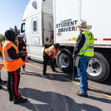CDL Training instructor stands by white semi truck with students in orange vests